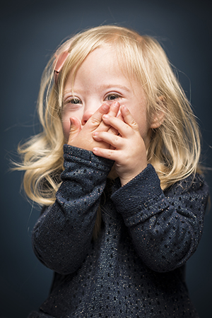 Porträttfoto i studio på Esther Isaksson, en liten flicka med kromosomavvikelse håller båda händerna mot munnen.