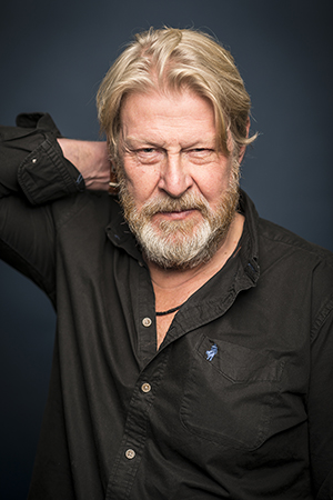 Porträttfoto i studio på Rolf Lassgård i svart skjorta med ena handen bakom nacken.