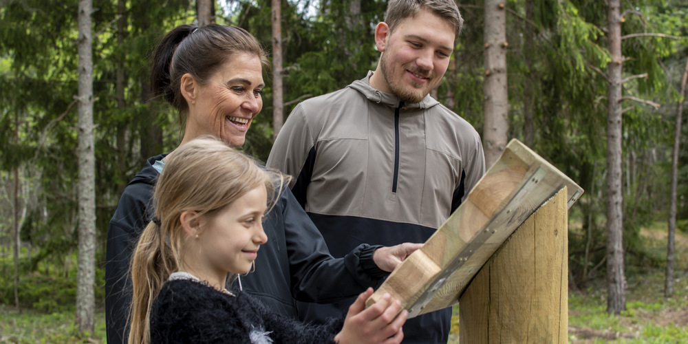 Ett barn, en kvinna och en man står och läser på en skylt i skogen