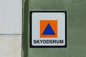 En skylt med vit bakgrund, en orange fyrkant med en blå triangel i och texten SKYDDSRUM.