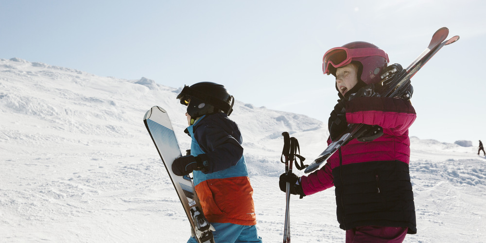 Två barn i en skidbacke. Båda bär sin utrustning. Ena barnet snowboard och andra barnet skidor.