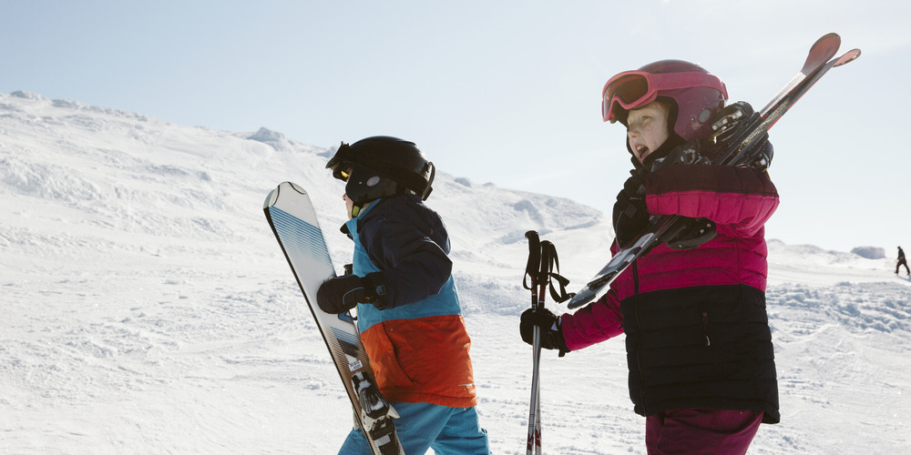 Två barn, ett bär på en snowboard och en på skidor i en skidbacke.